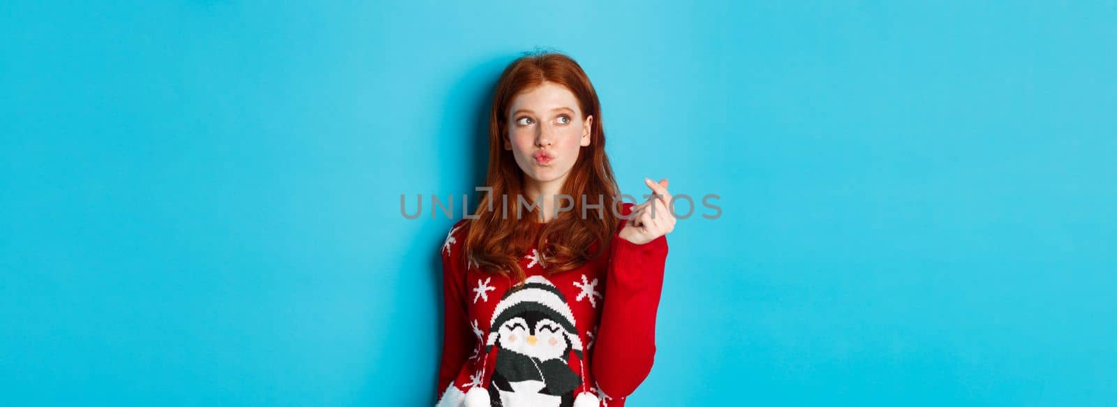 Winter holidays and Christmas Eve concept. Lovely redhead woman in xmas sweater, showing heart sign and thinking, looking upper left corner at logo, blue background.