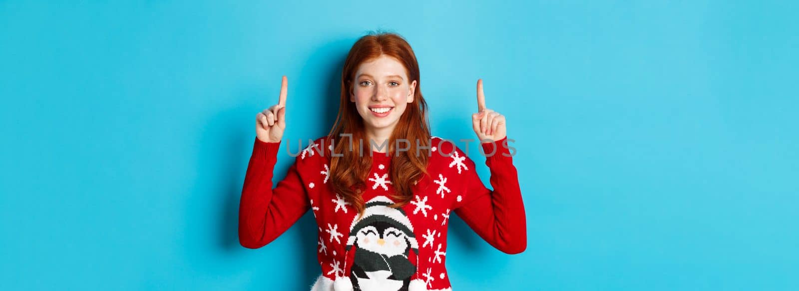 Winter holidays and celebration concept. Cute redhead girl in Christmas sweater, smiling and pointing fingers up at promo logo, standing over blue background.