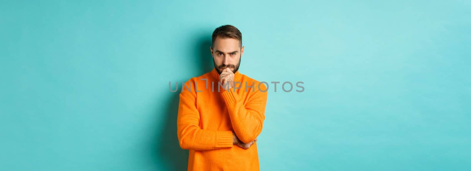 Thougtful young man making decision, looking serious and thinking, choosing, standing near copy space turquoise background.