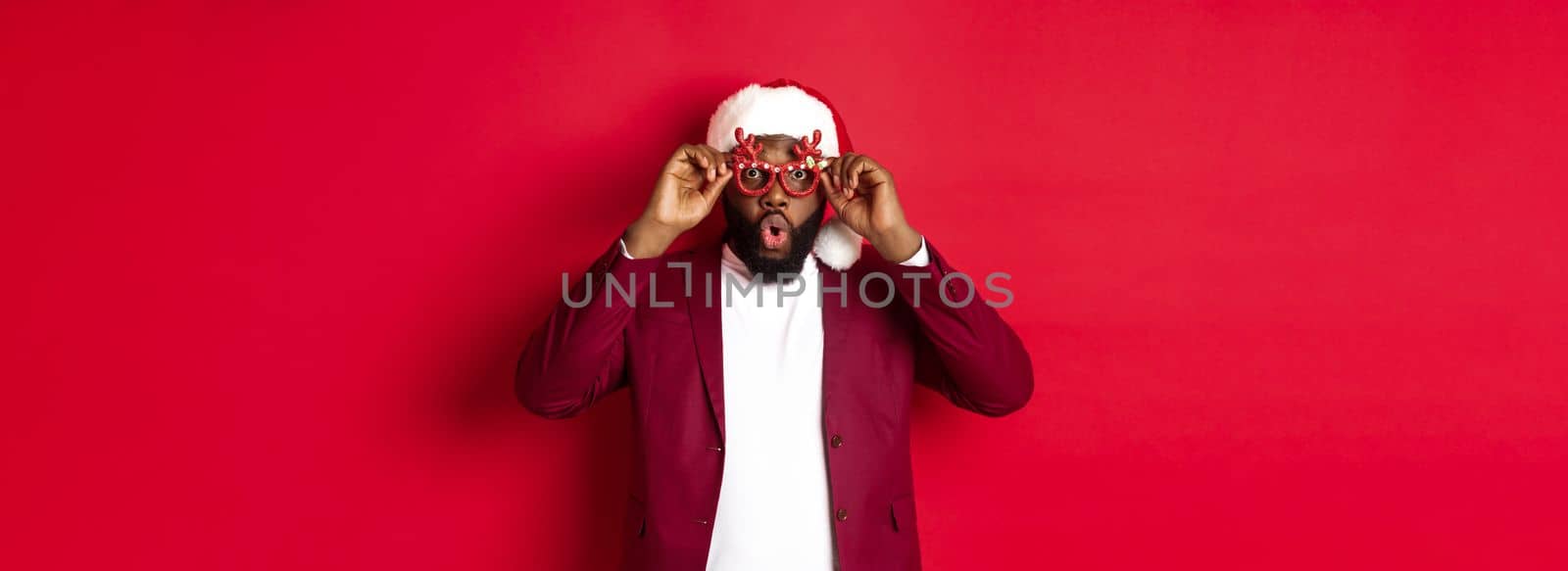 Funny Black man celebrating New Year, wearing party glasses and santa hat, having fun over red background.