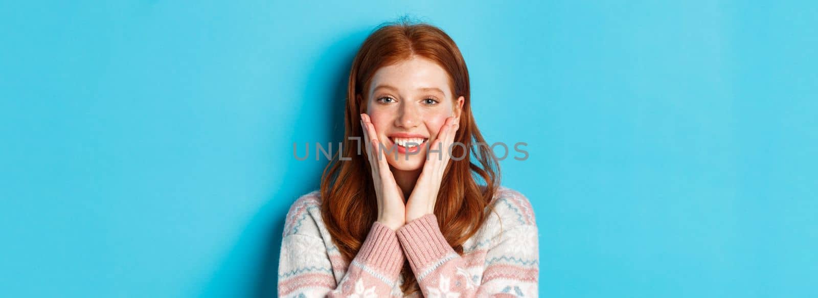Close-up of cheerful redhead girl blushing, touching cheeks and smiling happy, looking delighted at camera, standing over blue background.