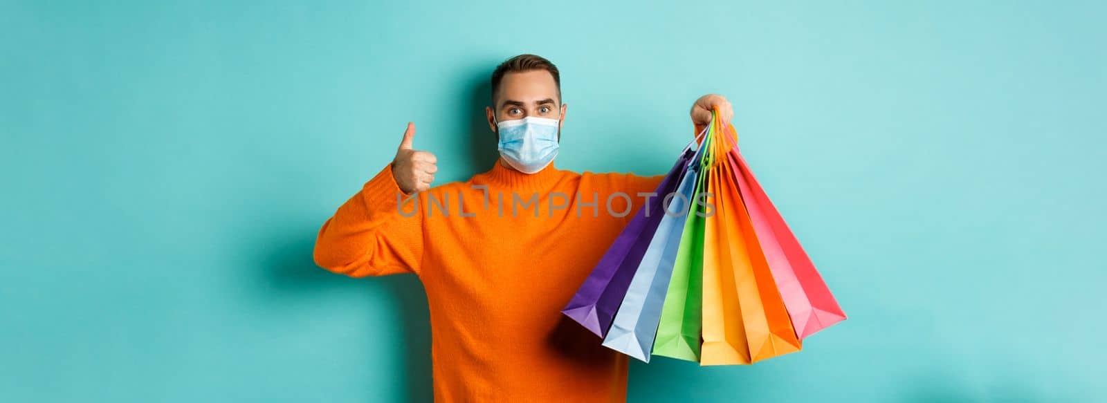 Covid-19, social distancing and lifestyle concept. Young man in face mask showing shopping bags and thumb-up, buying holiday gifts during pandemic.