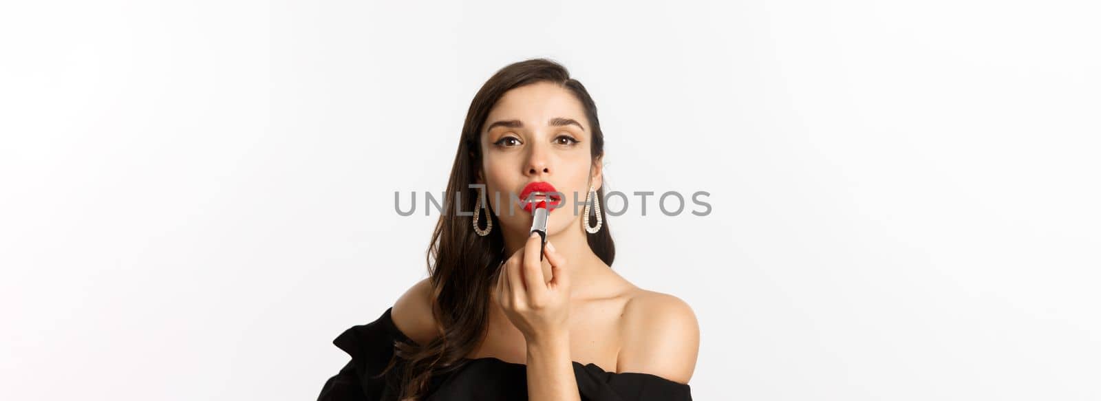 Close-up of beautiful woman applying red lipstick on lips, looking at camera like mirror, standing in black dress over white background by Benzoix