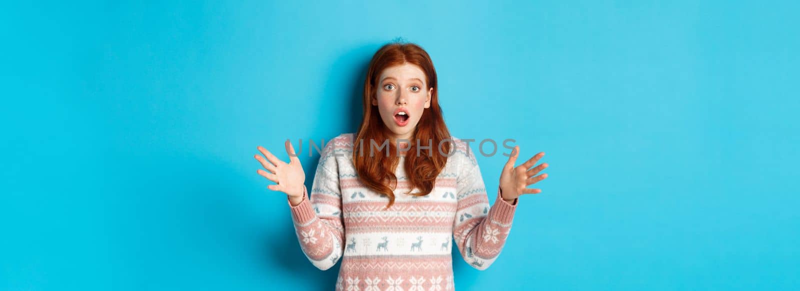 Image of startled redhead woman staring at camera with hands spread sideways, catching something, standing in winter sweater against blue background.
