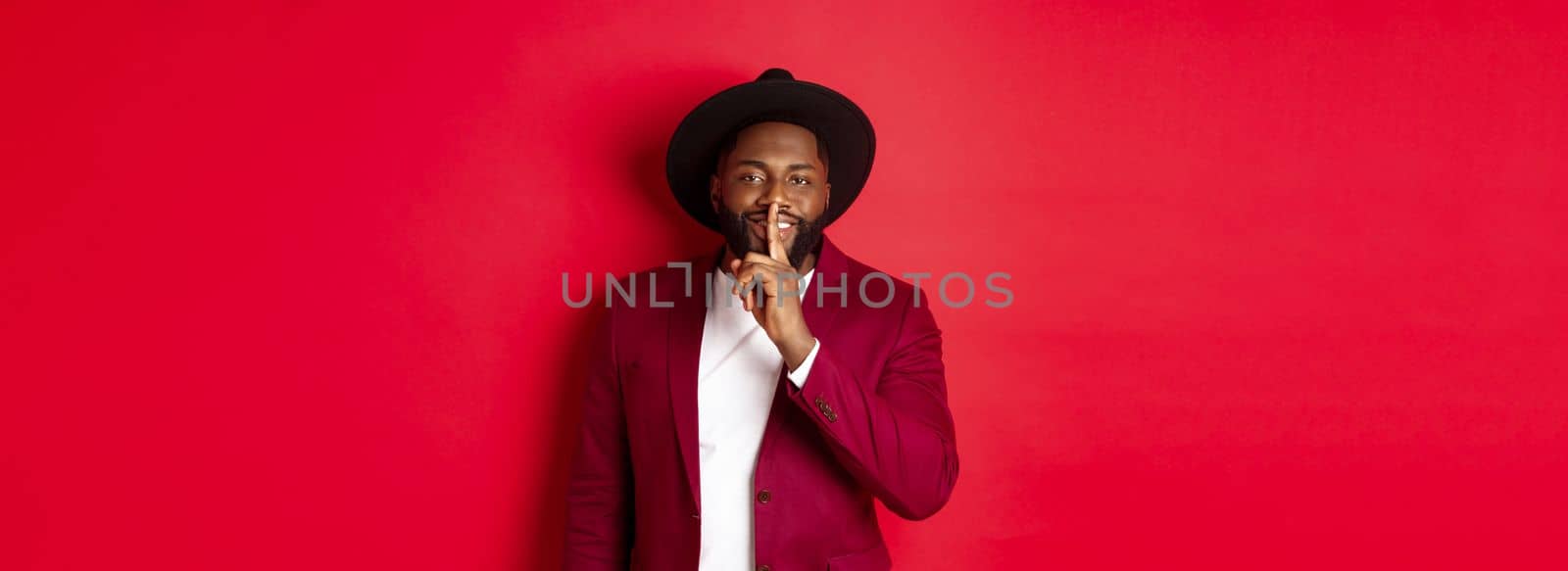 Winter holidays and shopping concept. Handsome Black man sharing secret, smiling and hushing at you with finger pressed to lips, standing against red background.