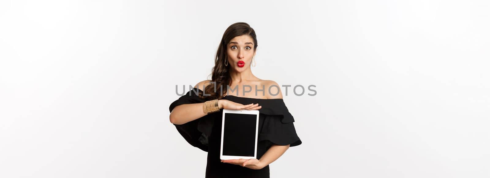 Fashion and shopping concept. Beautiful woman with red lipsticks, black dress, showing tablet screen and looking excited, standing over white background.