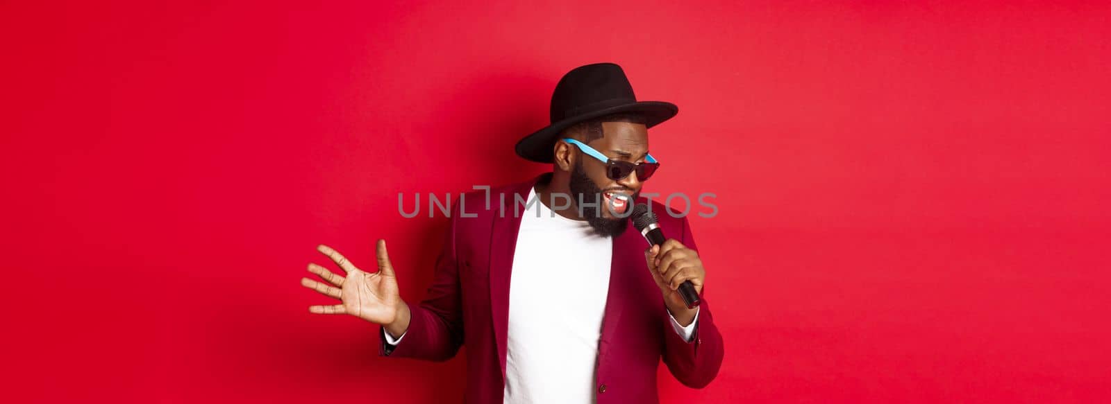 Passionate black male singer performing against red background, singing into microphone, wearing party outfit, standing over red background.