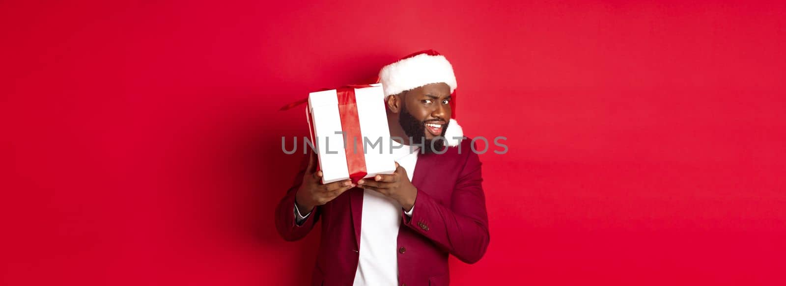 Christmas. Curious Black man in santa hat shaking new year gift, wonder what inside box, standing against red background.