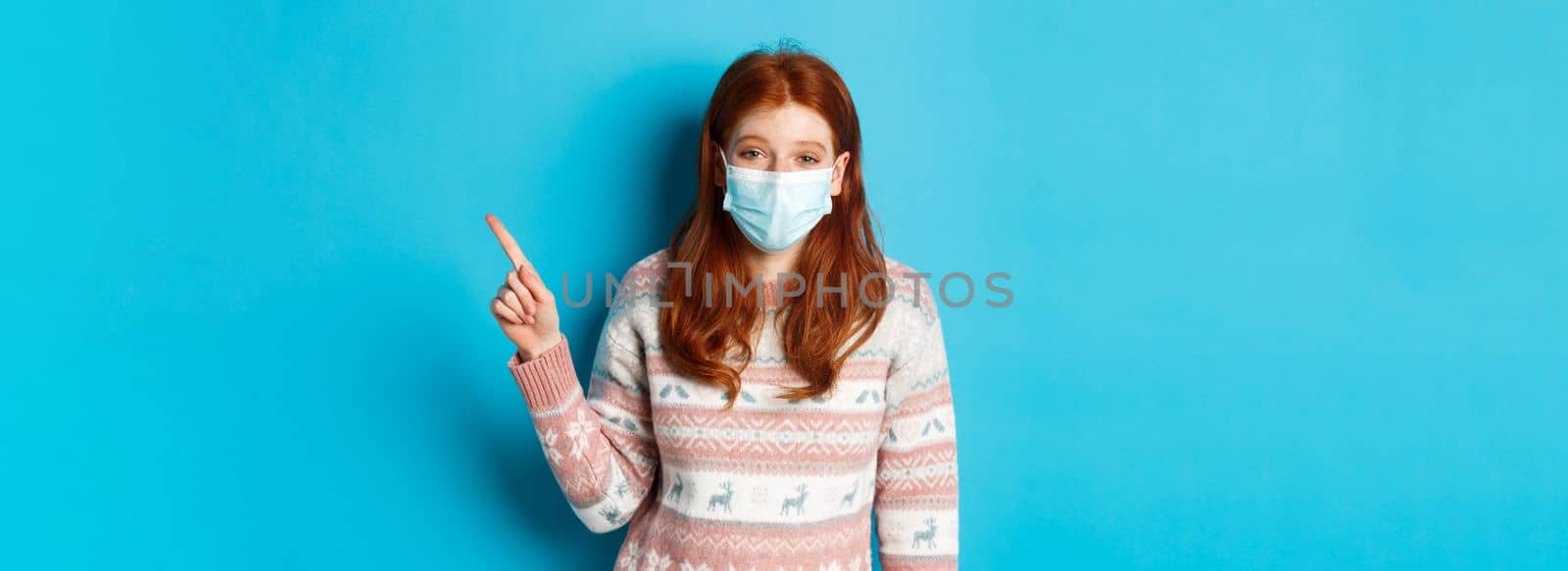 Winter, coronavirus and social distancing concept. Redhead girl in medical mask and sweater pointing at upper right corner and laughing, standing over blue background.