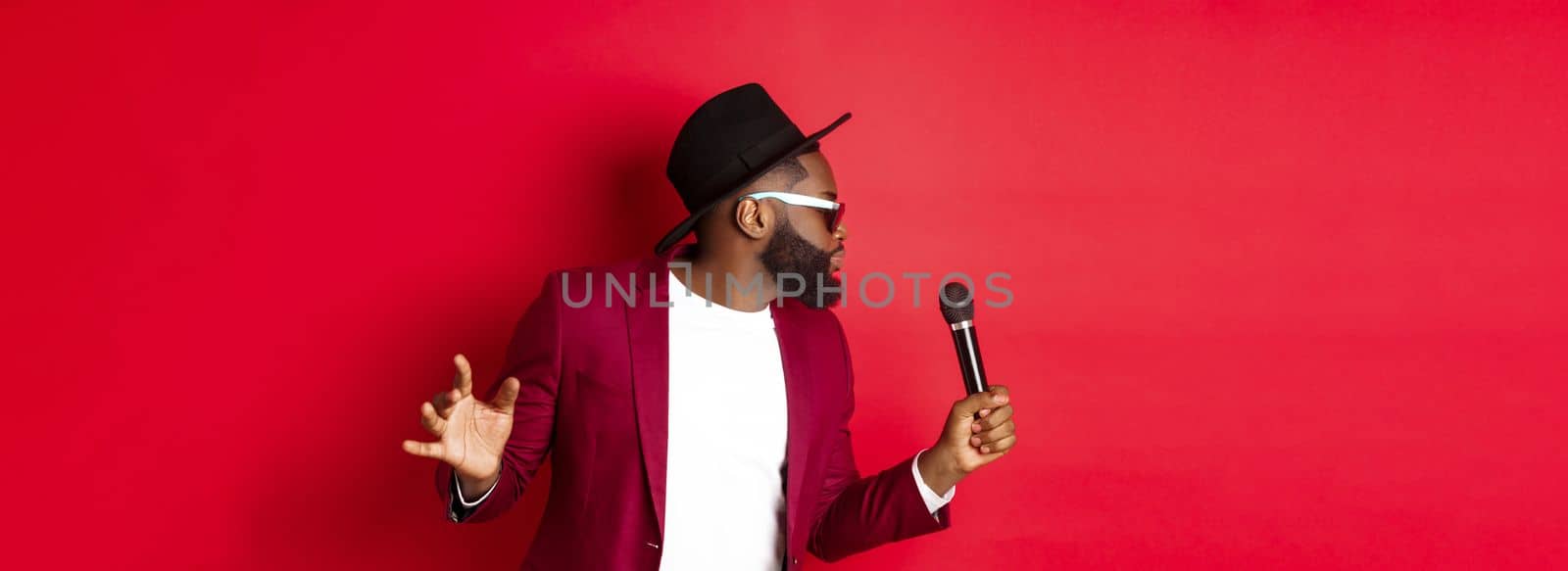 Passionate black male singer performing against red background, singing into microphone, wearing party outfit, standing over red background.