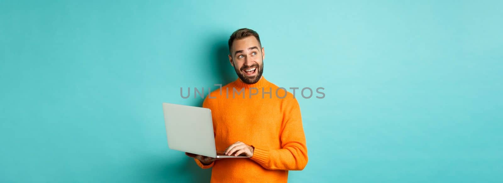 Handsome man doing online shopping, looking up thoughtful while using laptop, standing over light blue background. Copy space