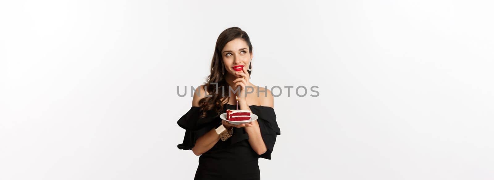 Celebration and party concept. Dreamy woman in black dress making wish, thinking and holding birthday cake with candle, standing over white background.