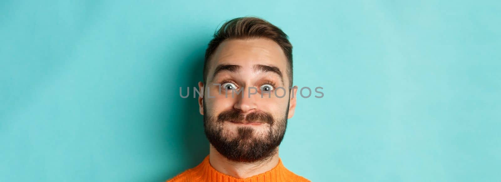 Headshot of funny bearded man showing faces and pouting, holding breath, standing in orange sweater against turquoise background.