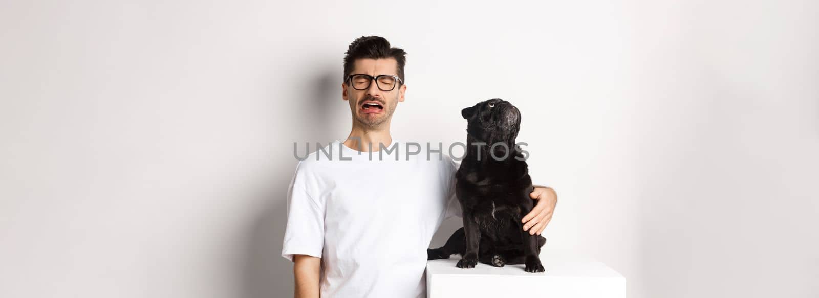Image of sad crying man hugging his cute black pug, dog looking curious at upper left corner promo logo, standing over white background.
