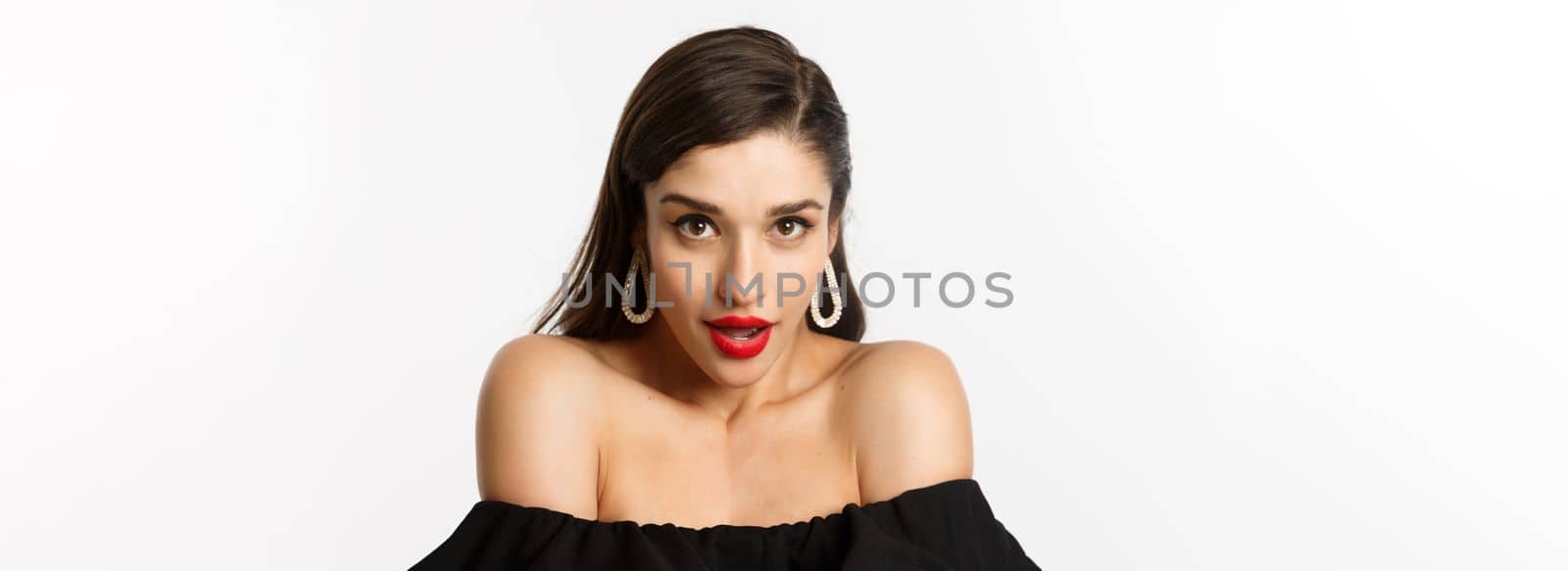Close-up of gorgeous brunette woman wearing elegant earrings and black dress, looking sensual at camera with piercing gaze and opened mouth, standing over white background.