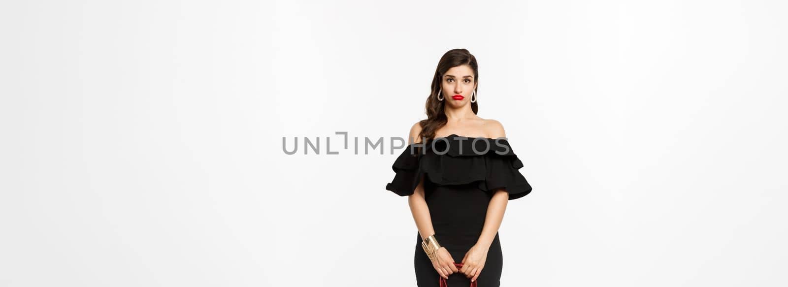 Beauty and fashion concept. Full length if silly young woman pouting and looking confused, holding purse, wearing heels and black dress, white background.