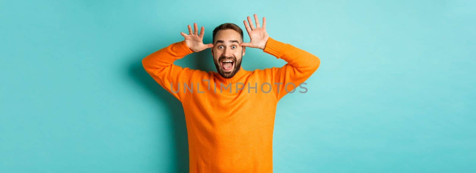 Image of handsome caucasian man making funny faces, mocking someone and smiling, standing against light blue background.