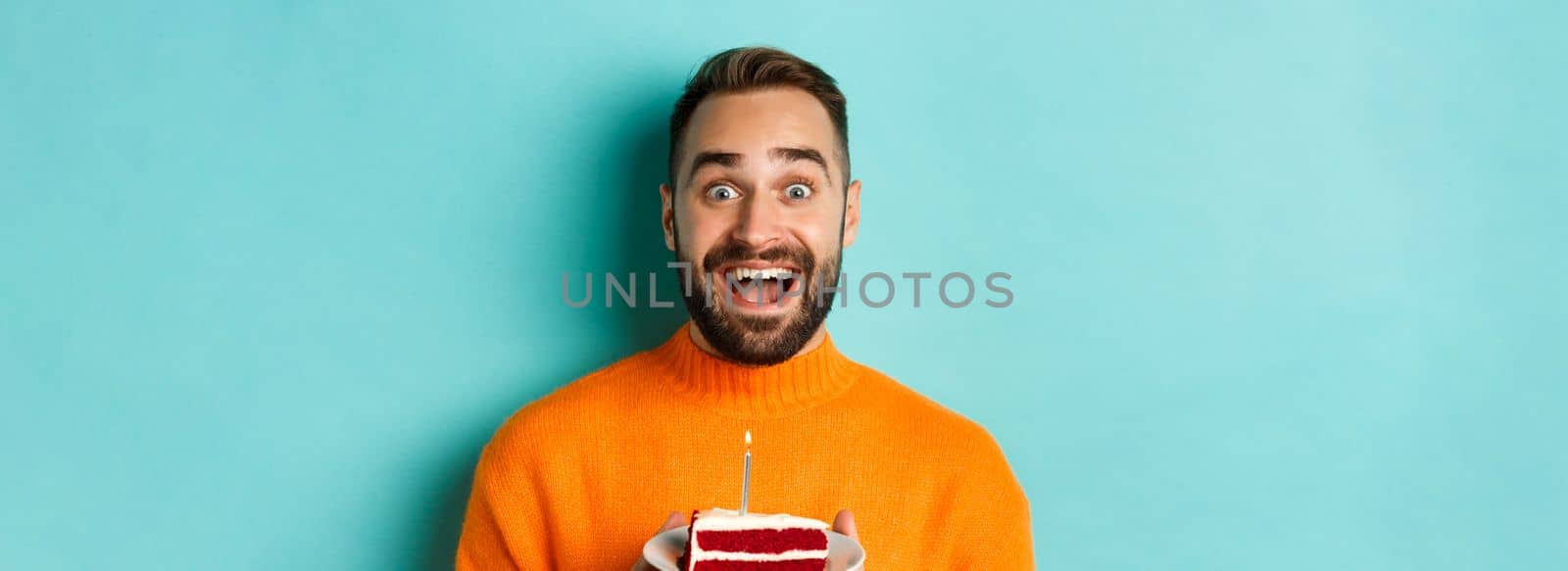 Close-up of happy adult man celebrating birthday, holding bday cake with candle and making wish, standing against turquoise background by Benzoix