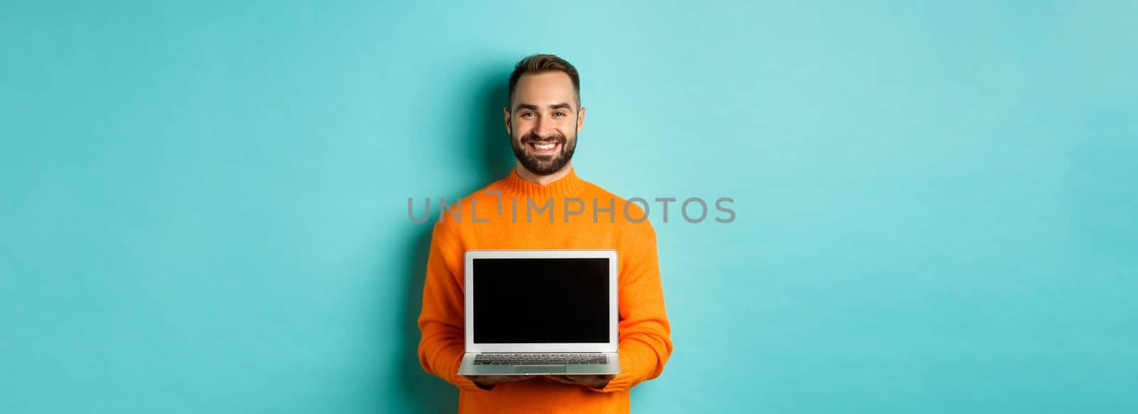 Handsome bearded man in orange sweater showing laptop screen, demonstrating online store, standing over light blue background by Benzoix