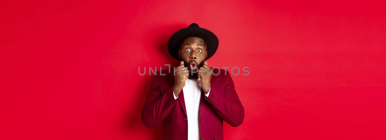 Fashion and party concept. Surprised african american male model looking shocked and amazed, saying wow and staring camera, standing against red background.