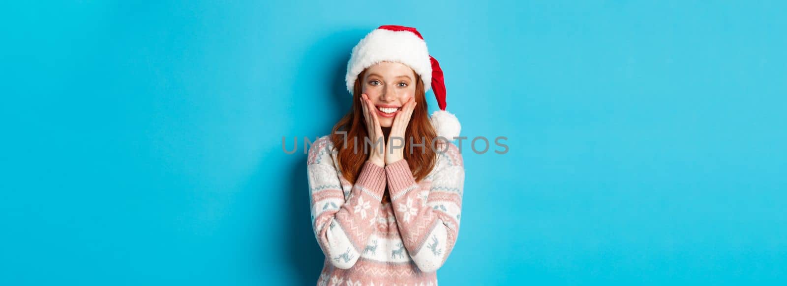 Winter and Christmas Eve concept. Happy redhead girl celebrating xmas, staring at camera amazed, smiling and touching cheeks, standing over blue background.