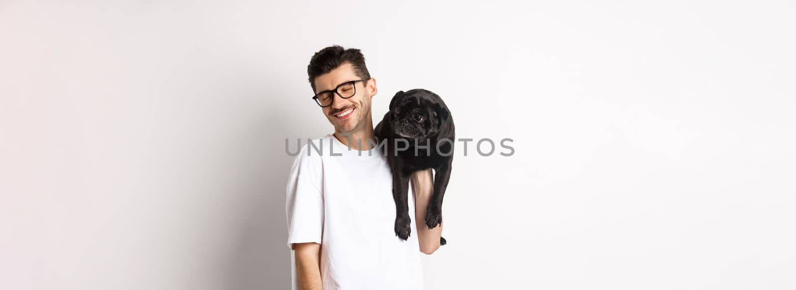 Happy man in love with dog, holding puppy on shoulder and smiling, standing over white background.