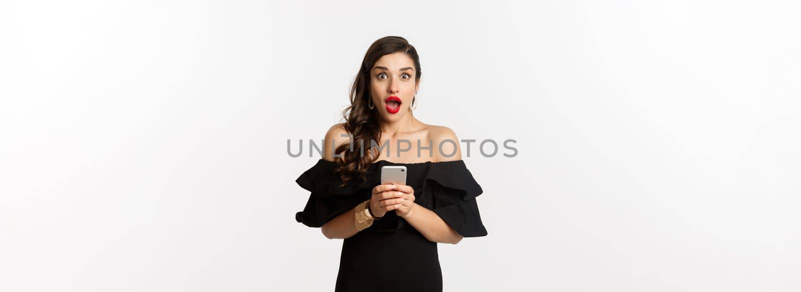 Happy young glamour woman in black dress, holding mobile phone and looking surprised, standing over white background.