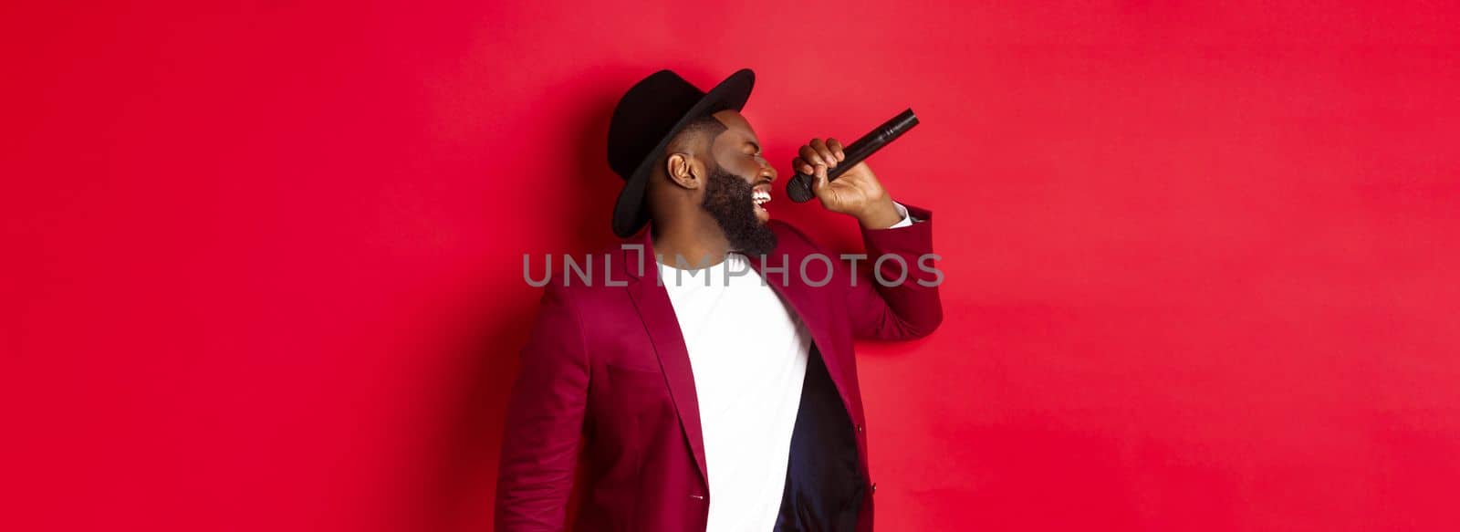 Passionate black male singer performing against red background, singing into microphone, wearing party outfit, standing over red background.