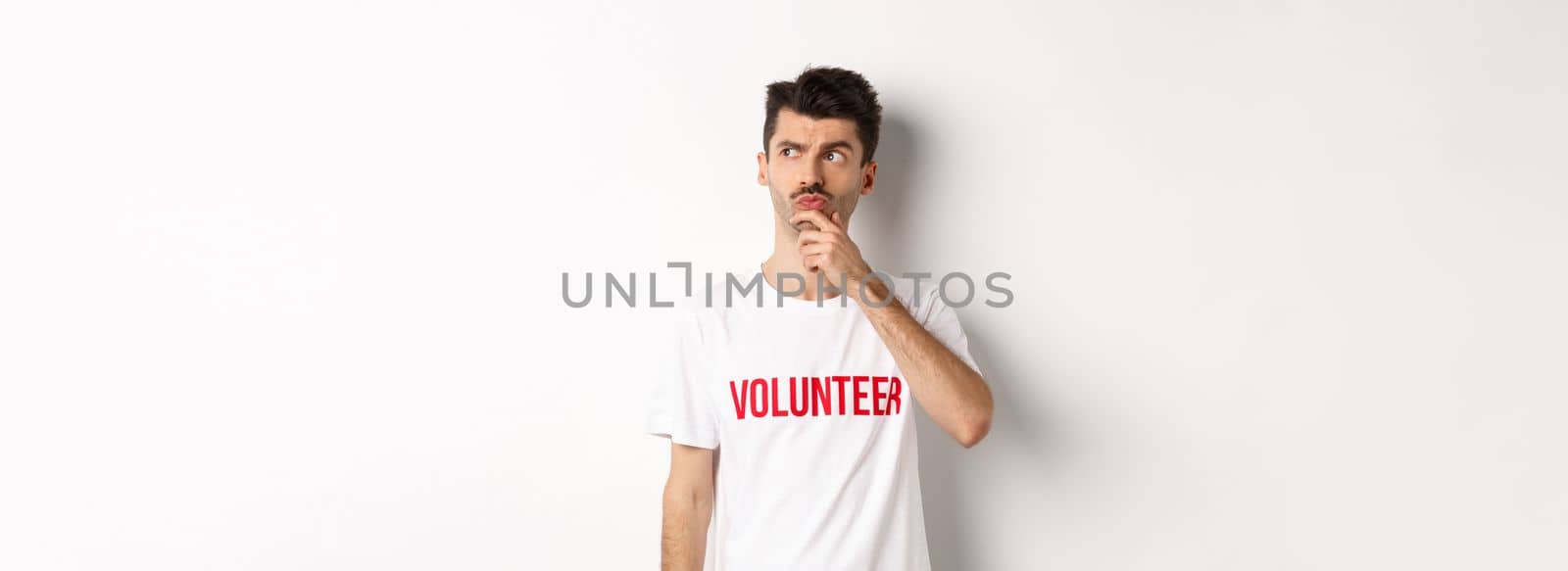Thoughtful young male volunteer in white t-shirt searching solutions, looking at upper left corner and thinking, standing over studio background.