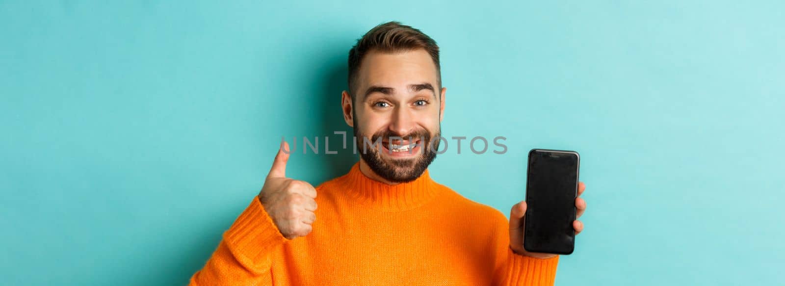 Close-up of attractive bearded man showing smartphone screen, thumbs up, recommending mobile app, standing satisfied over turquoise background by Benzoix