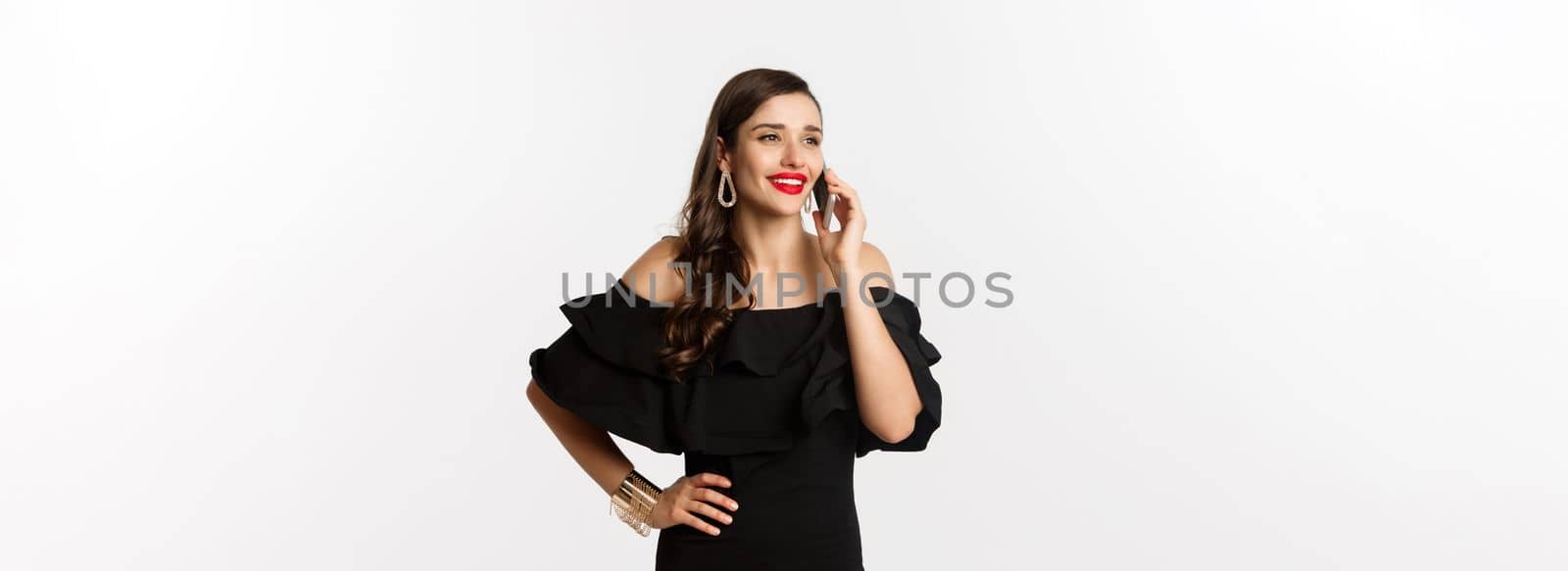 Successful young woman in black dress, red lipstick and makeup, talking on mobile phone and smiling, standing over white background by Benzoix