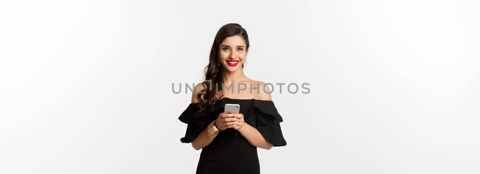 Online shopping concept. Attractive young woman in black dress, reading text message, using mobile phone and smiling, standing over white background by Benzoix