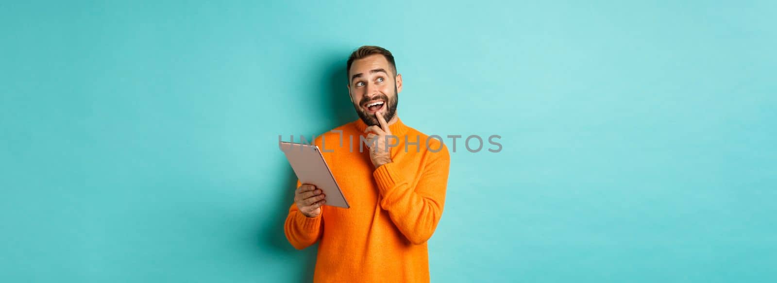 Handsome man thinking and using digital tablet, shopping online and pondering ideas, looking at upper left corner, standing over light blue background.