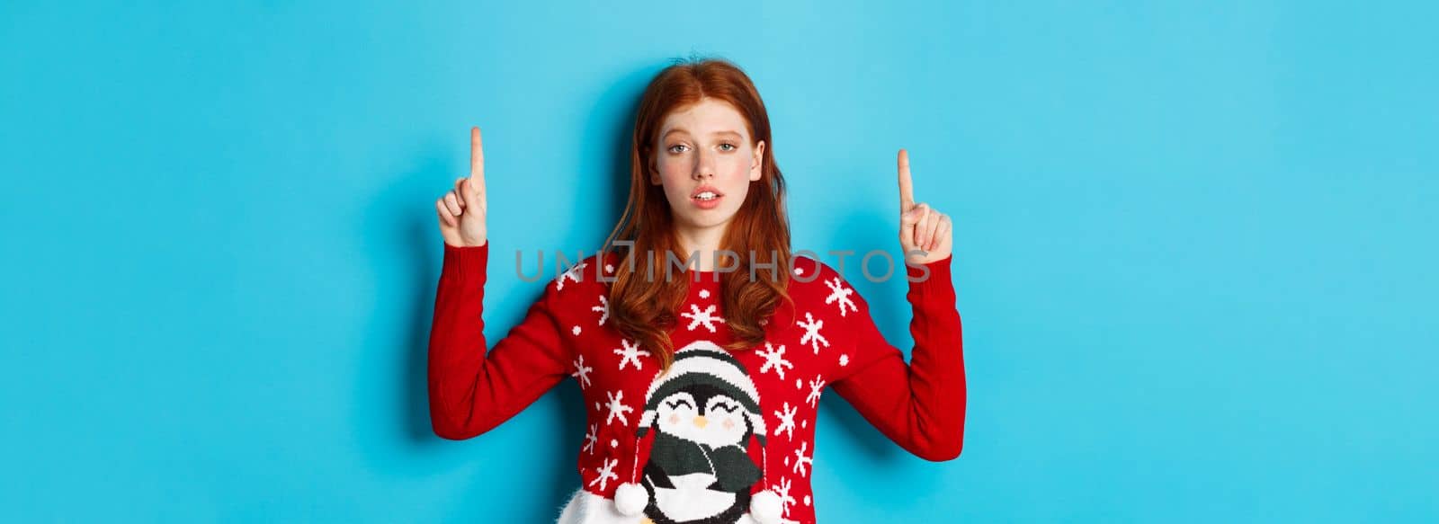 Merry Christmas. Skeptical and unamused redhead girl pointing fingers up, showing logo with reluctant face, standing over blue background by Benzoix