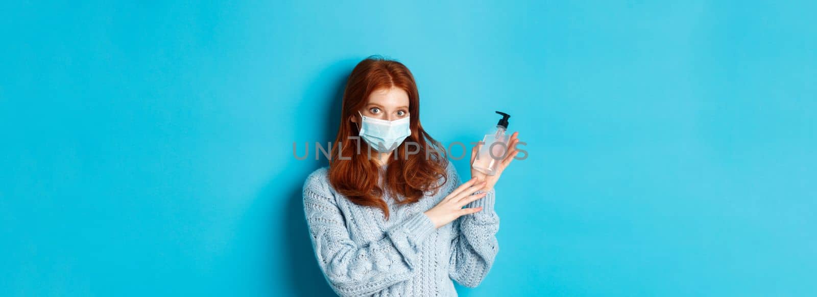Winter, covid-19 and social distancing concept. Young redhead girl in face mask showing hand sanitizer, demonstrating antiseptic for disinfection, standing over blue background.