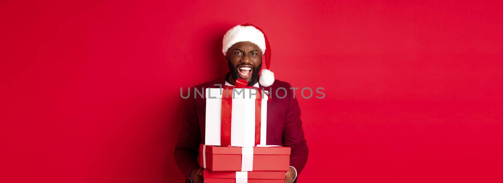 Christmas, New Year and shopping concept. Happy Black man in santa hat and blazer holding xmas presents, bring gifts and smiling, standing against red background.