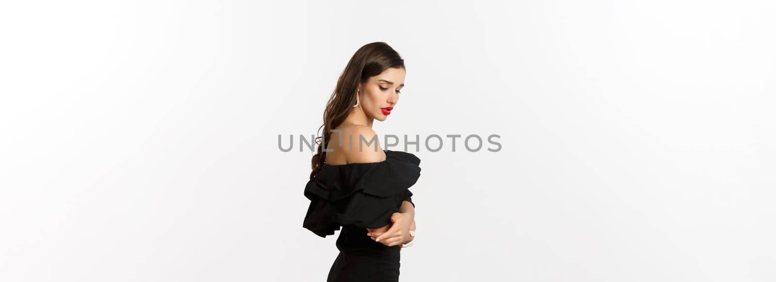 Profile of sensual brunette woman in black elegant dress, looking down, standing over white background.