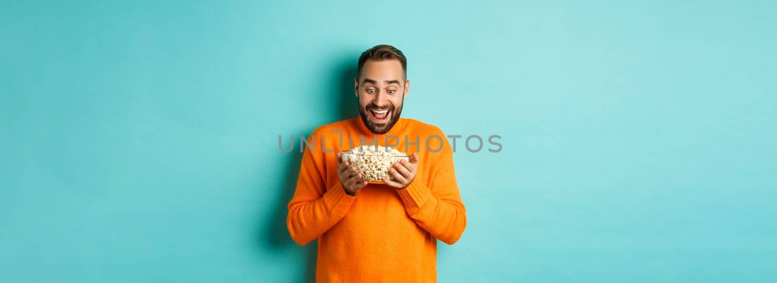 Handsome bearded man looking with exctied and happy at popcorn bowl, ready to watch movie, standing over white background.