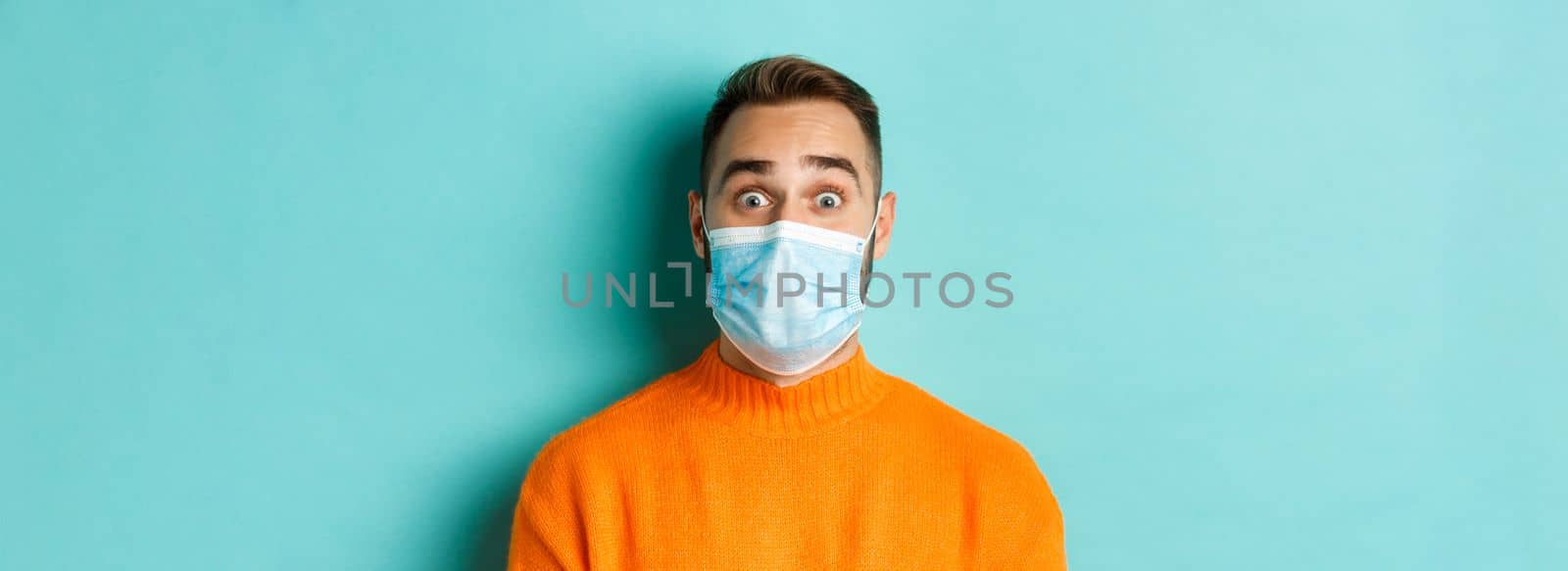 Covid-19, social distancing and quarantine concept. Close-up of young man in face mask staring at camera with surprised face, standing over turquoise background.