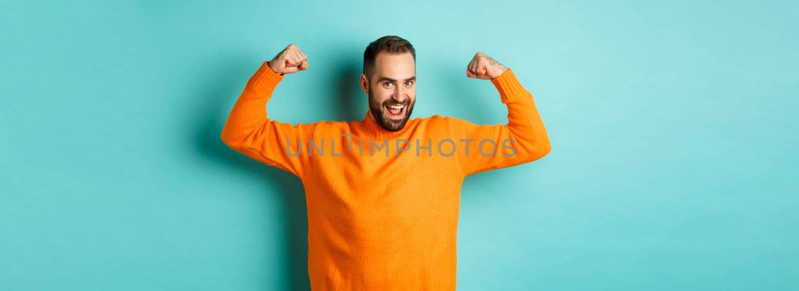 Young and strong man looking confident, flex biceps and show-off muscle strengths, smiling self-assured, standing over light blue background by Benzoix