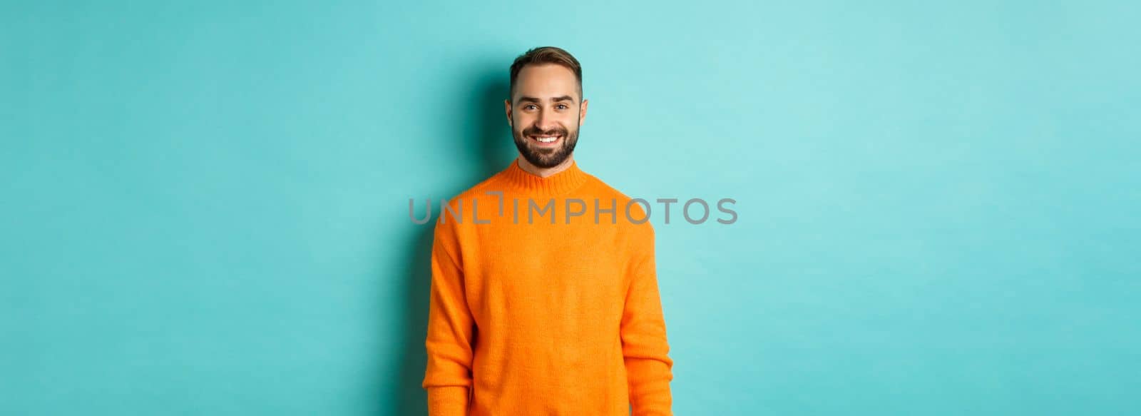 Lifestyle. Handsome man in orange sweater smiling at camera, happy face, guy cheerful expression, standing over turquoise background by Benzoix