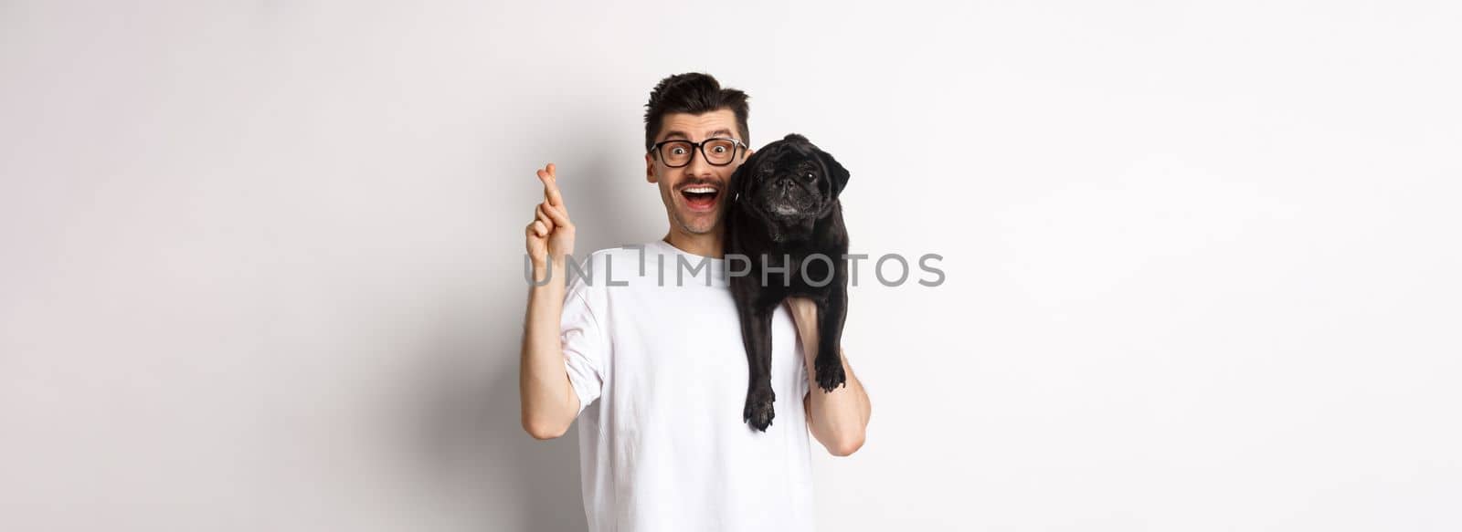 Hopeful smiling dog owner making a wish, holding cute black pug on shoulder and cross fingers for good luck, white background.