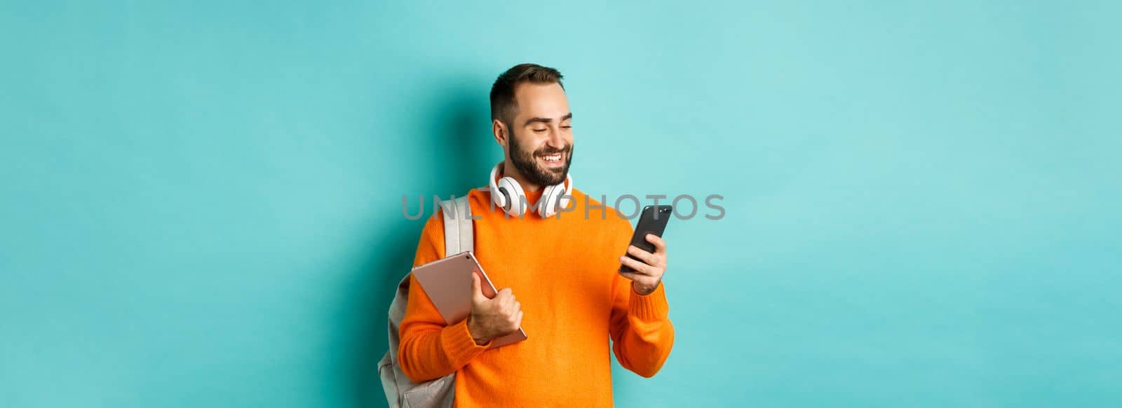 Handsome man student with headphones and backpack, holding digital tablet, reading message on mobile phone, standing against light blue background.