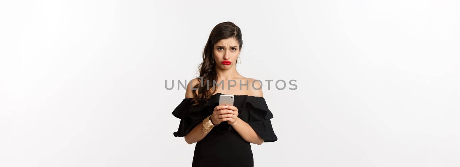 Sad and gloomy woman in black dress, sulking upset, using mobile phone and feeling disappointed, standing over white background.