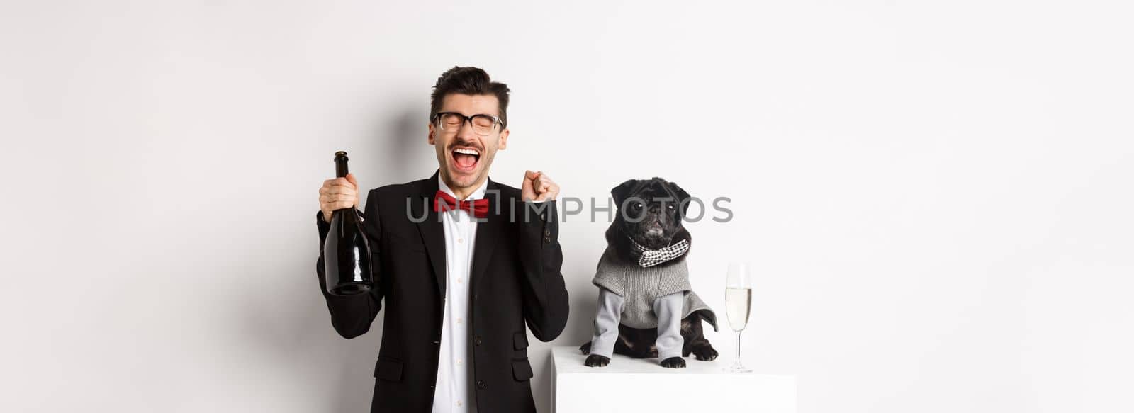 Pets, winter holidays and New Year concept. Happy young man celebrating Christmas with cute black dog wearing party costume, holding bottle champagne, white background.