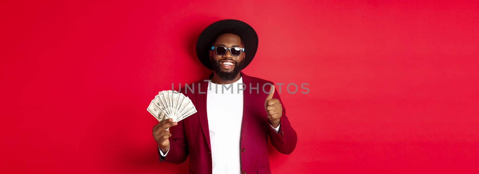 Handsome modern african american man in sunglasses and party clothes, showing thumb up with dollars, earn money and looking satisfied, red background.