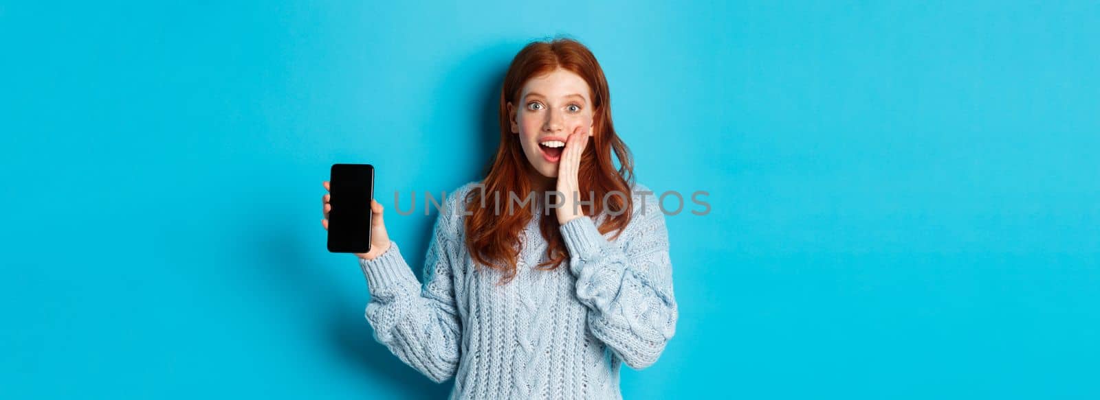 Amazed redhead girl looking at camera, showing smartphone screen, demonstrating mobile application, standing over blue background by Benzoix