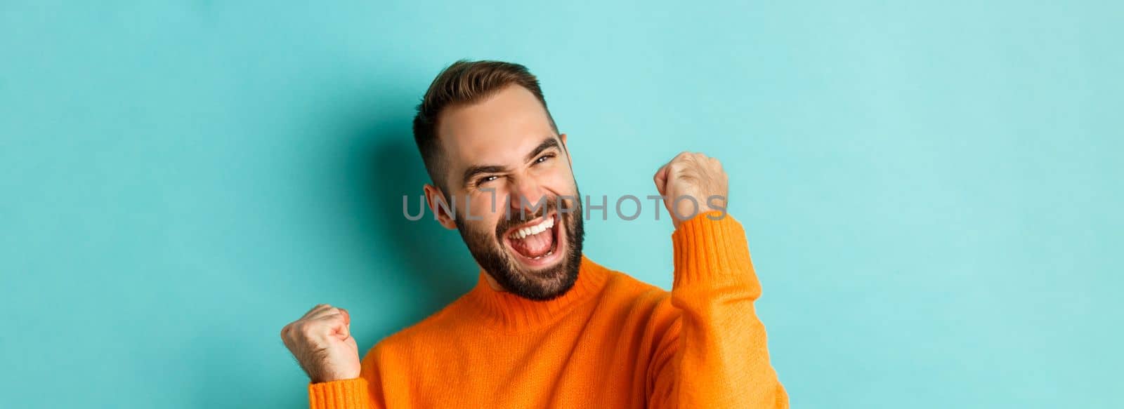 Image of handsome relieved man feeling satisfaction, rejoicing of winning or achievement, making fist pump and saying yes, standing over turquoise background.