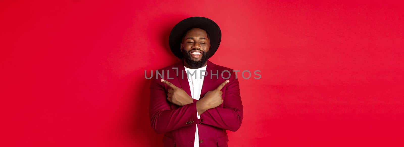 Happy young man dreaming and pointing fingers sideways, close eyes and smiling, showing two things, standing over red background.