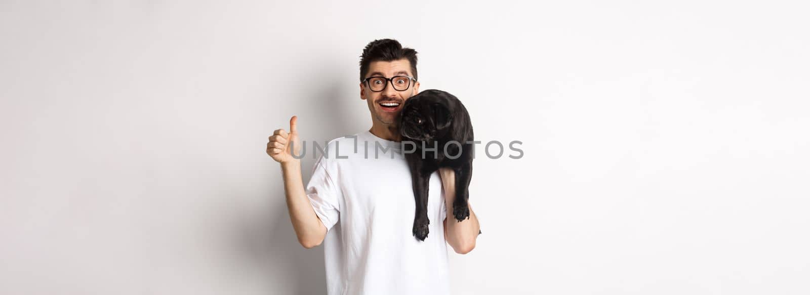 Happy and satisfied dog owner showing thumb-up, holding cute black pug on shoulder, recommending pet products, standing over white background.